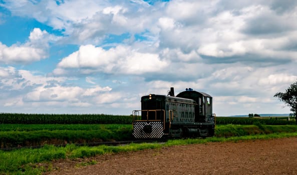 A vintage locomotive moves along railway tracks surrounded by lush green fields, with fluffy clouds overhead.