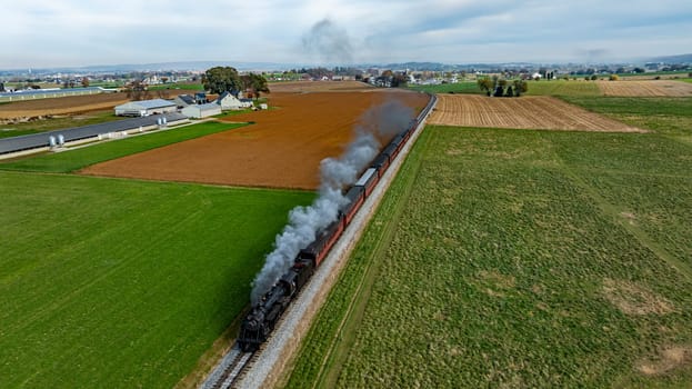 A steam train moves along the tracks surrounded by green fields and farmland under a cloudy sky.