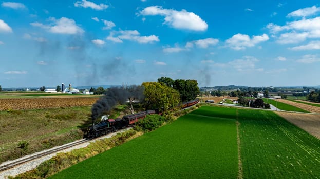 A steam train billows black smoke as it moves through vibrant green fields and trees under a clear blue sky.