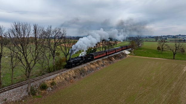 Ronks, Pennsylvania, USA, October 4, 2023 - A steam locomotive puffs clouds of smoke as it moves along a track through fields and trees under a cloudy sky.