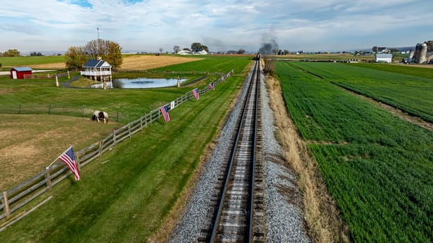 A train moves through a countryside setting lined with American flags, featuring a pond and lush fields in autumn.