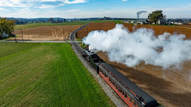 A vintage steam train puffs white smoke as it moves through green and brown fields under a clear blue sky.