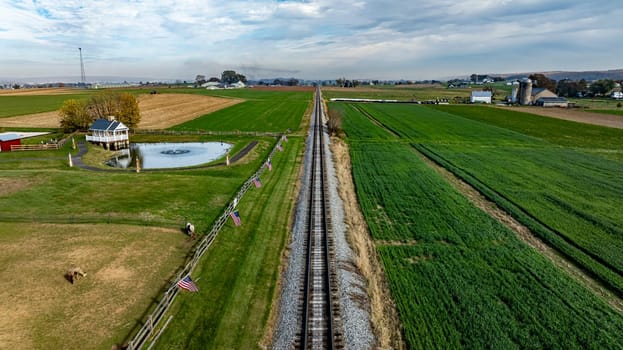 A serene rural landscape features railroad tracks leading through green fields and farms under a cloudy sky.