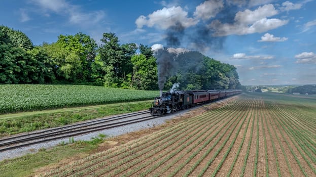 Ronks, Pennsylvania, USA, June 8, 2024 - A Black Steam Locomotive Billowing Thick Smoke Pulls Red Passenger Cars Through Lush Green Fields And Forested Areas, Under A Clear Blue Sky With Puffy Clouds, Creating A Picturesque Countryside Journey.