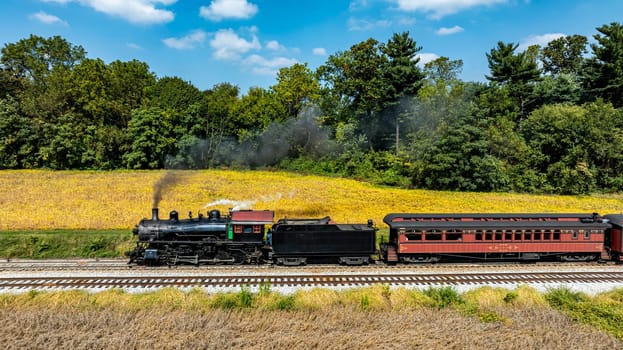 A steam train chugs along the railway, passing vibrant golden fields under a clear blue sky.