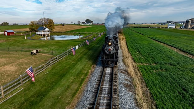 A steam train chugs along a railway in a green countryside, passing by flags and fields under a clear sky.