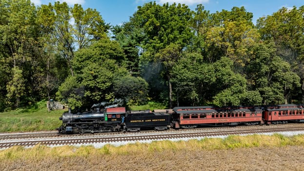 Ronks, Pennsylvania, USA, October 4, 2023 - A vintage steam locomotive pulls a passenger car along tracks surrounded by vibrant trees under a clear sky.