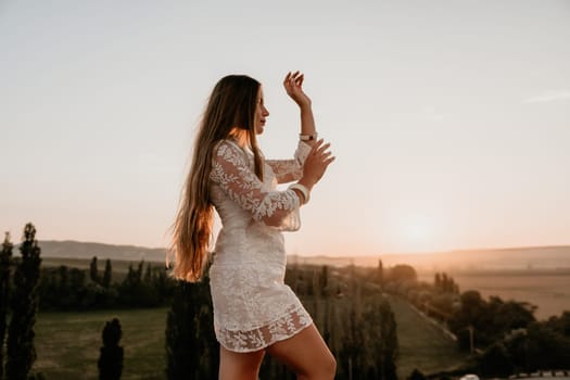 Romantic beautiful bride in white dress posing with sea and mountains in background. Stylish bride standing back on beautiful landscape of sea and mountains on sunset