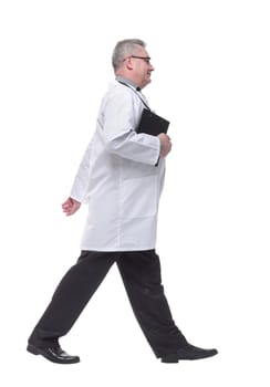 Full length profile shot of a male doctor in a white uniform walking isolated on white background with clipboard and stethoscope