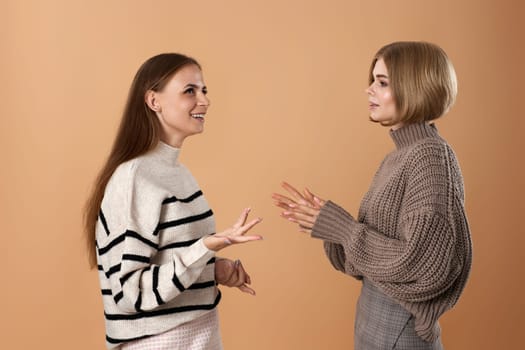 Two cheerful attractive women friends talking together while standing on beige background