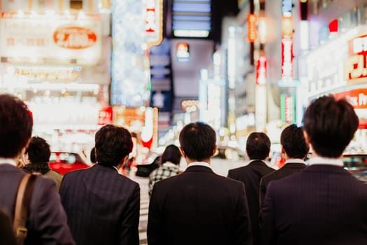 Japanese corporative business people in suits, waiting in rush hour on crossroad in Shinjuku business district, Tokyo, Japan. Blured advertising boards illuminated in the background.