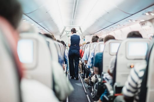 Interior of airplane with passengers on seats and stewardess in uniform walking the aisle.