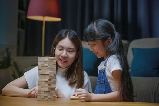 Asian young mother playing game in wood block with little daughter in home living room at night, Smiling woman help teach preschooler kid play build constructor tower of wooden blocks, family funny
