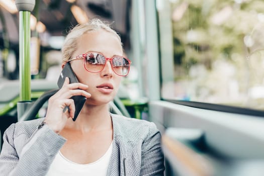 Blonde caucasian business woman talking on cell phone, traveling by bus. Public transport and commuting to work.