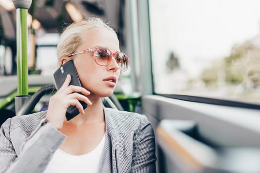 Blonde caucasian business woman talking on cell phone, traveling by bus. Public transport and commuting to work.