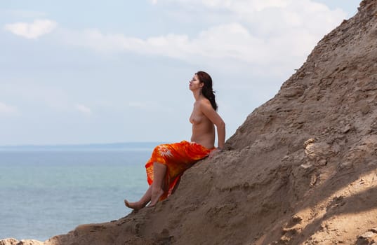 A young woman poses nude on a sand hill against a sky and seascape backdrop. Nature, beauty and nudity concept