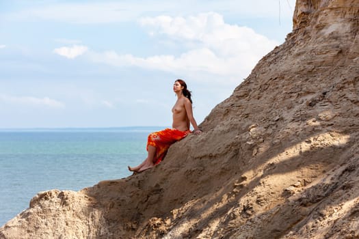 A young woman poses nude on a sand hill against a sky and seascape backdrop. Nature, beauty and nudity concept