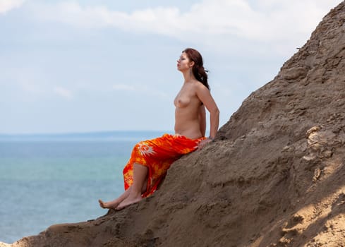A young woman poses nude on a sand hill against a sky and seascape backdrop. Nature, beauty and nudity concept