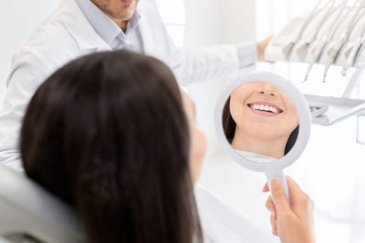 Happy female patient looking at mirror and checking her teeth in dental clinic, healthy smile concept, cropped