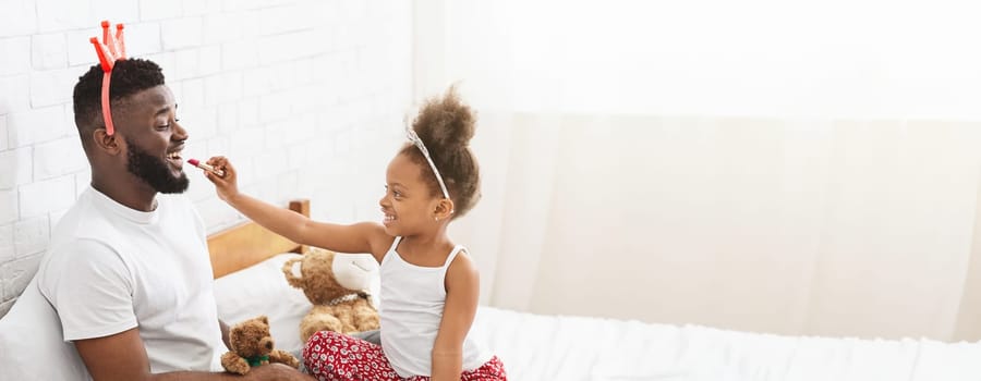 African American father and his daughter share joyful laughter as they engage in playful interaction on a bed filled with stuffed animals. The little girl playfully applies lipstick on her father lips