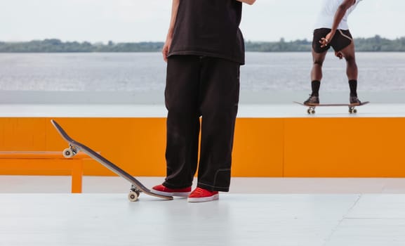 Man's legs with skateboard in skatepark. Professional skater athlete in black jeans holding skateboard with his foot. Another skateboarder rides a skateboard in the background at a skate park