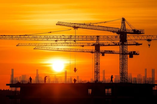 Silhouettes of cranes and construction workers against a vibrant orange sunset sky, symbolizing urban development.