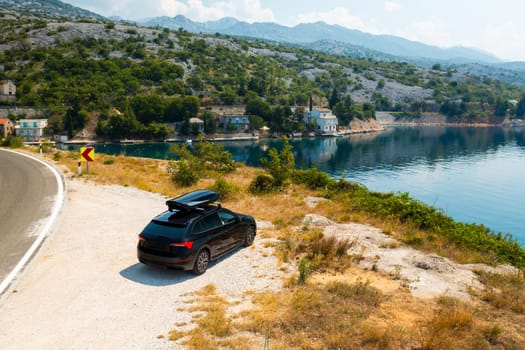Black car with a roof box, parked at a viewpoint on a high cliff, overlooking a tranquil, azure sea
