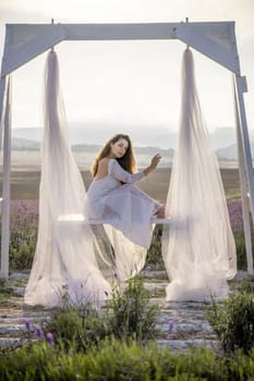 A woman is sitting on a bench under a white canopy. She is wearing a white dress and has long hair. The scene is set in a field with purple flowers