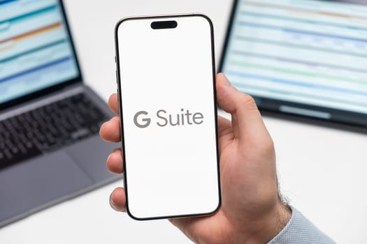 PRAGUE, CZECHIA - JULY, 2024: Google Suite logo on the screen of smartphone in mans hand and laptop on the background