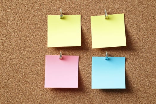 Four blank sticky notes in yellow, pink, and blue colors pinned in a grid pattern on cork board.