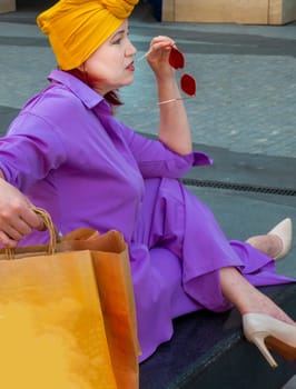 woman with craft shopping bags in the city. The concept of fashionable Consumerism and ecology. An adult Asian woman in a purple suit with red and yellow accents enjoys shopping . a sunny day.