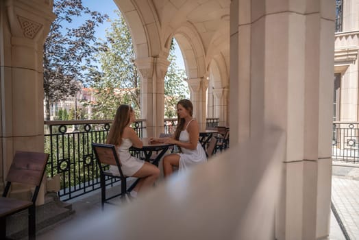 Two women in white dresses sit at a table on a balcony, chatting. The balcony has an ornate railing and is surrounded by arched columns.