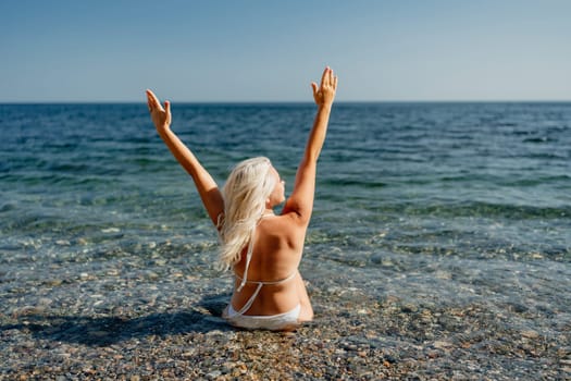 A woman is sitting in the ocean, with her arms raised in the air. She is wearing a bikini and she is enjoying the water