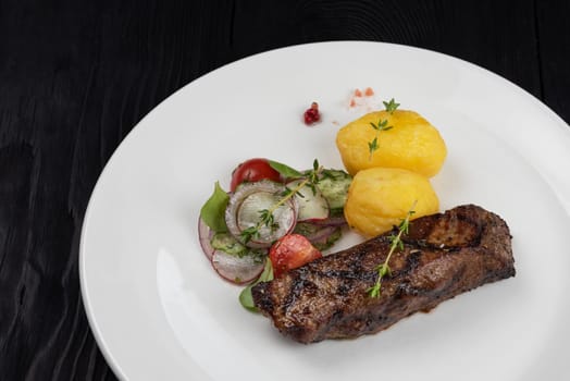 Grilled beef skirt steak meat with potato and vegetables on white plate on wooden black background
