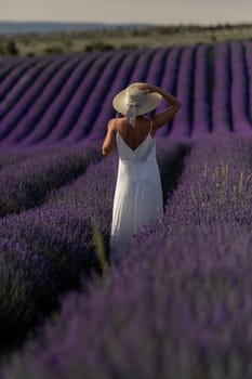 A woman in a white dress is walking through a field of purple flowers. She is wearing a straw hat and she is enjoying the scenery