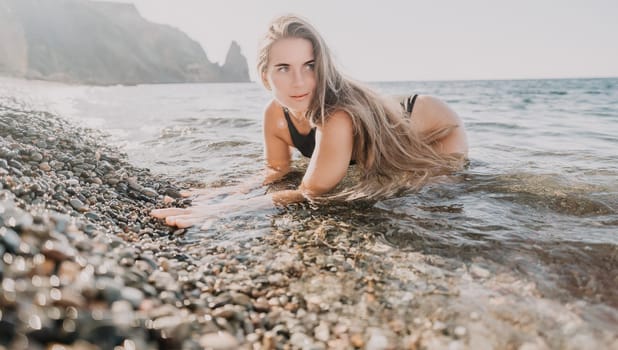 A young woman in a black bikini lies in the shallow water of a pebble beach, looking off to the side.