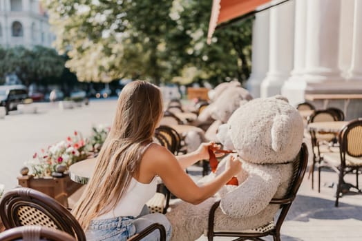 A woman sits cafe with a teddy bear next to her. The scene is set in a city with several chairs and tables around her. The woman is enjoying her time at the outdoor cafe