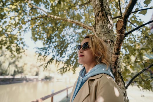 A blonde woman in sunglasses leans against a tree with a serene lake in the background.