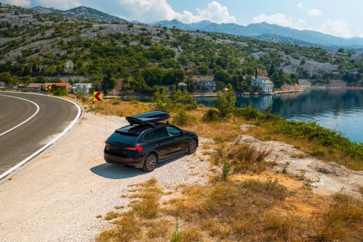 Black car with a roof box parked near a winding road on a high cliff, with the azure sea sparkling under the bright sunlight