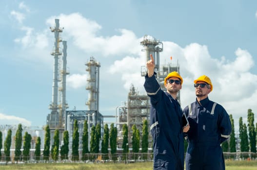 Two factory engineer or technician workers with one hold tablet and point forward look like explain work to coworker and stand in front of refining petrochemical factory with day light.