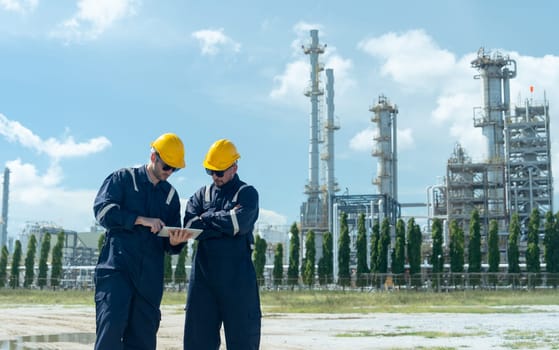 Wide shot of two factory engineer or technician workers discuss together with tablet and stand in front of refining and petrochemical factory with day light.