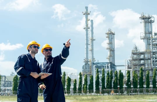 Two factory engineer or technician workers with one hold tablet and other point to right side and stand in front of refining petrochemical factory with day light.