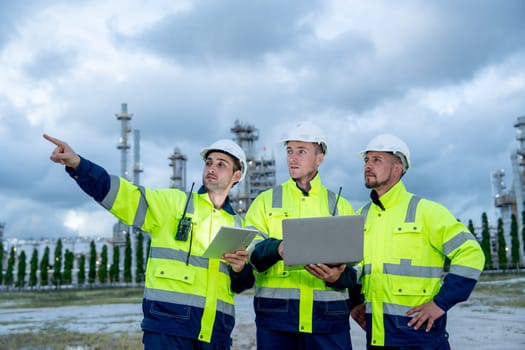 Group of engineer or technician workers stand in front of  petrochemical factory and use laptop to discuss their work with one point to left side.