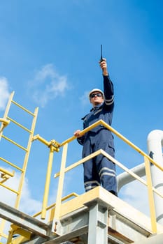 Vertical image factory engineer or technician worker man use walkie talkie to point forward and stand on high state over pipeline of petrochemical gas in workplace with blue sky as background.
