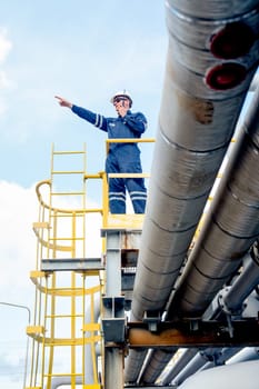 Vertical image of factory engineer or technician worker man use walkie-talkie to contact co-worker also point forward with stand on high state over pipeline of petrochemical gas in workplace.