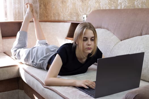 Young girl works at laptop while lying on sofa on her stomach in domestic room. Online office work from home, work of the future from your home room using computer