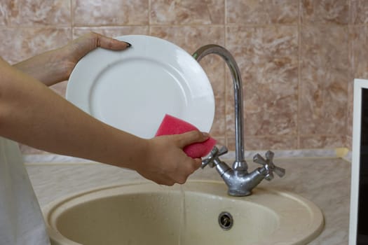 White plate in hands of housewife while washing dishes in the sink in kitchen after dinner, dish sponge in foam washes away the dirt on the plate