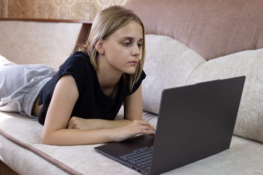 Young woman uses laptop while lying on sofa on her stomach, computer technology for working and studying from home, distance learning and performing work duties