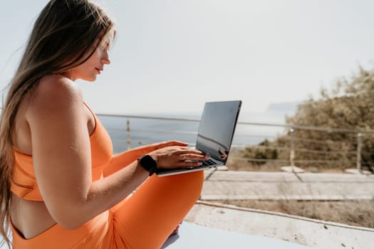 A young woman wearing orange sportswear sits on a yoga mat and uses her laptop computer outdoors with a blue sea in the background.