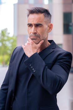 Close-up of a caucasian businessman looking serious at the camera in a suit standing with hand on chin with thoughtful face,in an outdoor workspace. Front view of a young lawyer with thoughtful expression looking